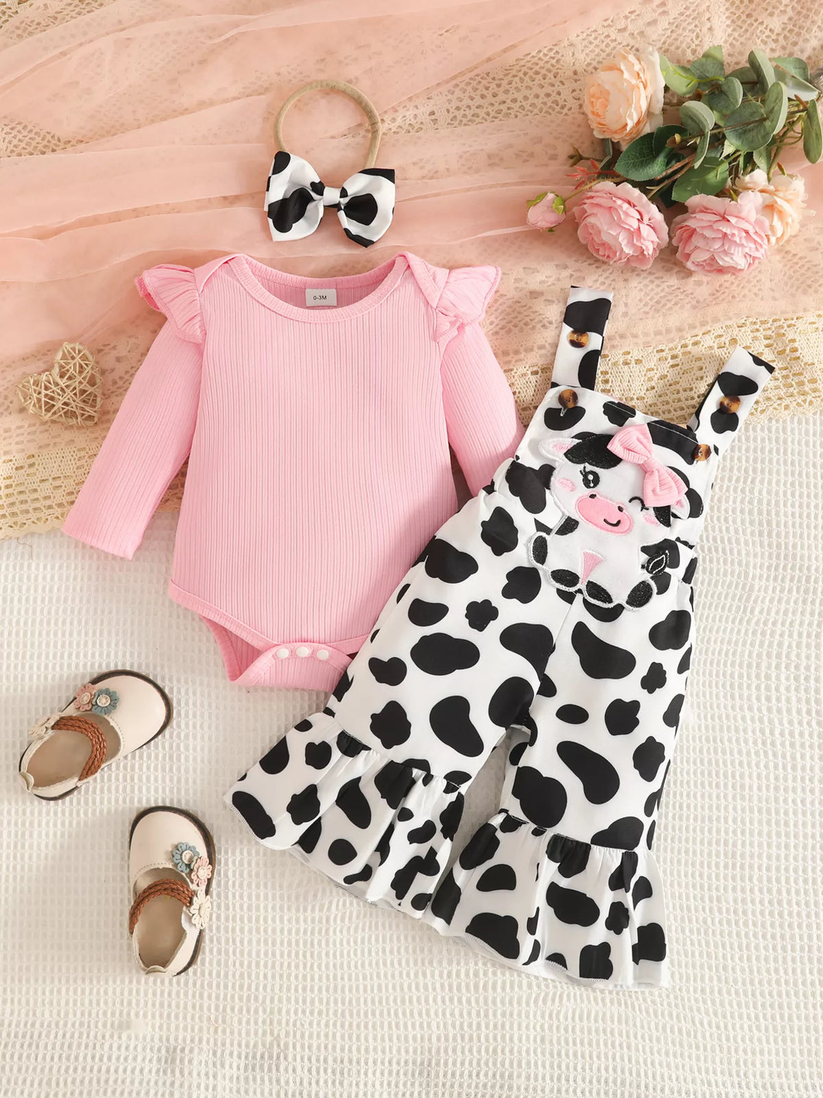 Pink baby bodysuit with black-and-white cow-print flared overalls and matching bow headband.