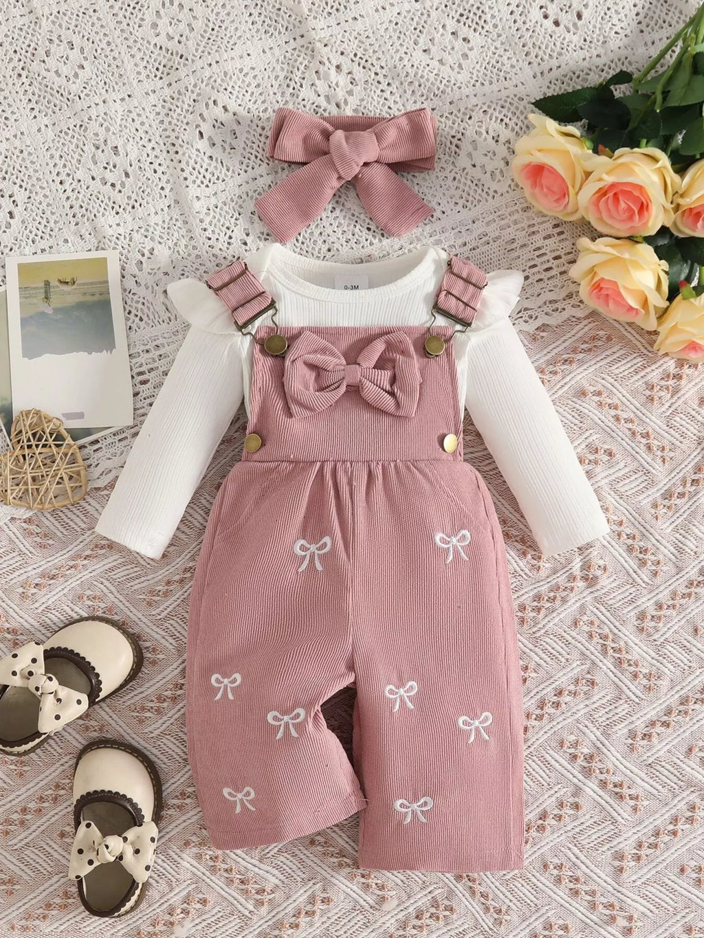 White ribbed baby bodysuit with pink bow-embroidered overalls and matching headband displayed flat