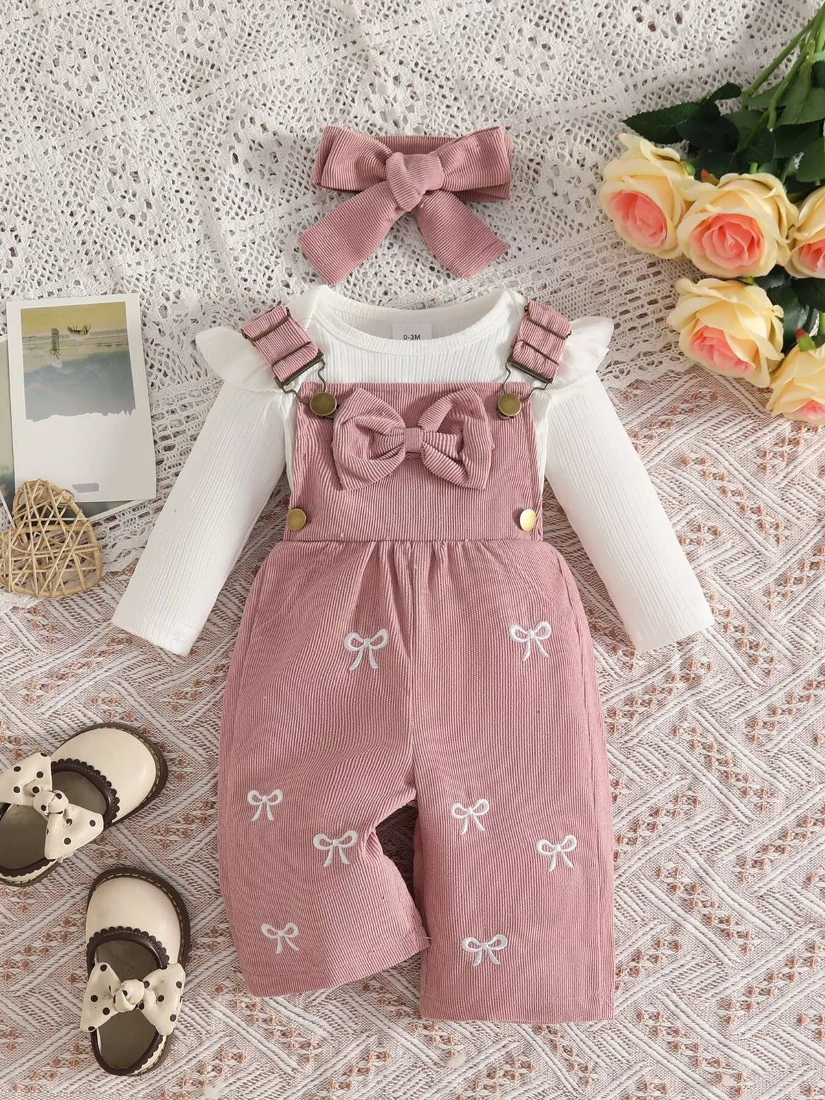 White ribbed baby bodysuit with pink bow-embroidered overalls and matching headband displayed flat