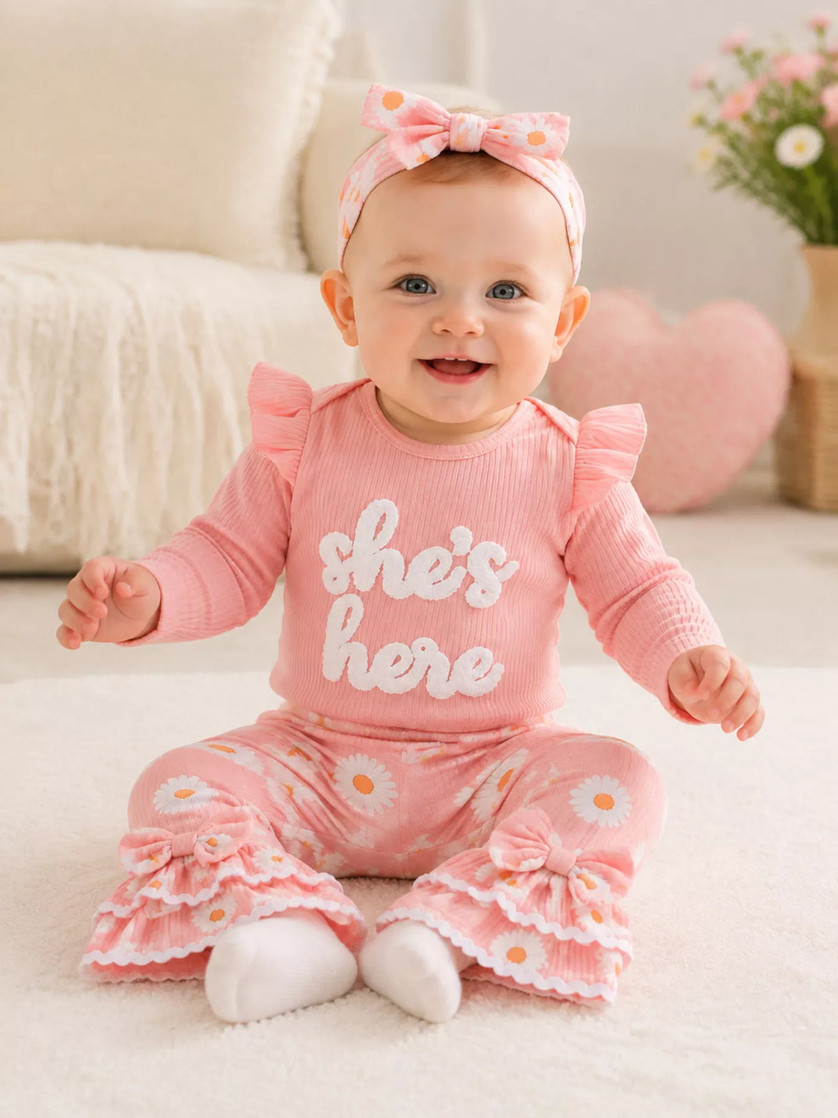 Baby wearing a pink She's Here ruffle-shoulder bodysuit, daisy-print flared pants and bow headband.