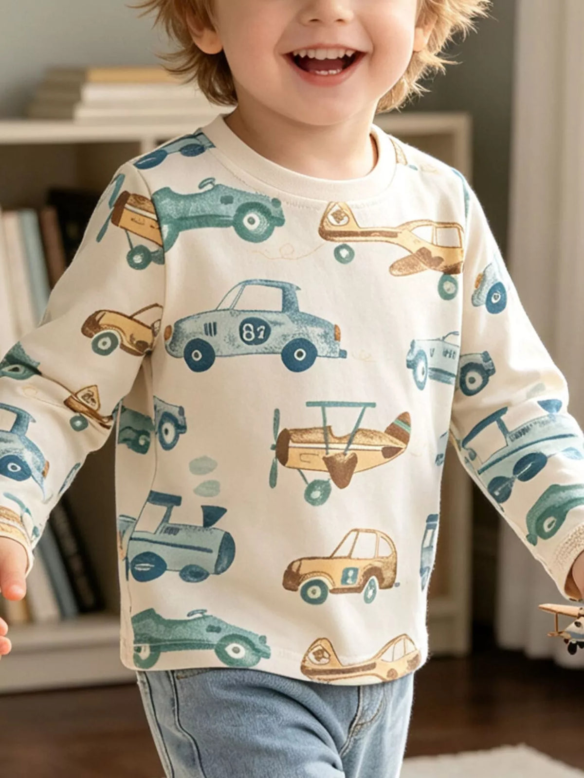 Child wearing a cream long-sleeve top printed with blue cars and tan airplanes.