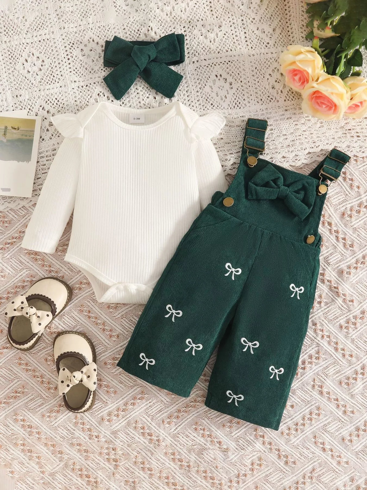 White ribbed baby bodysuit with dark green bow-embroidered overalls and matching headband displayed flat