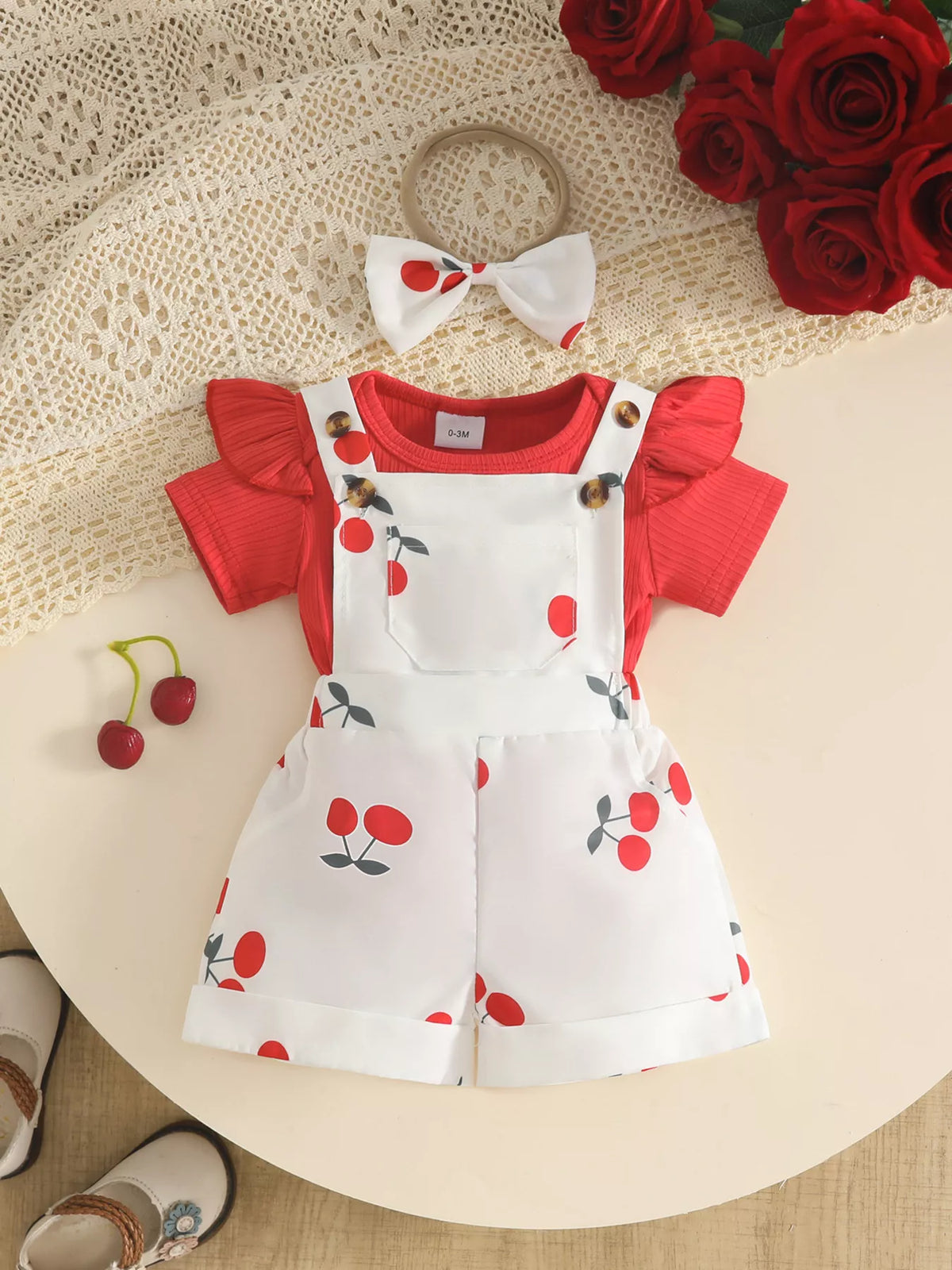Red ruffle-sleeve baby bodysuit with white cherry-print short overalls and matching bow headband.
