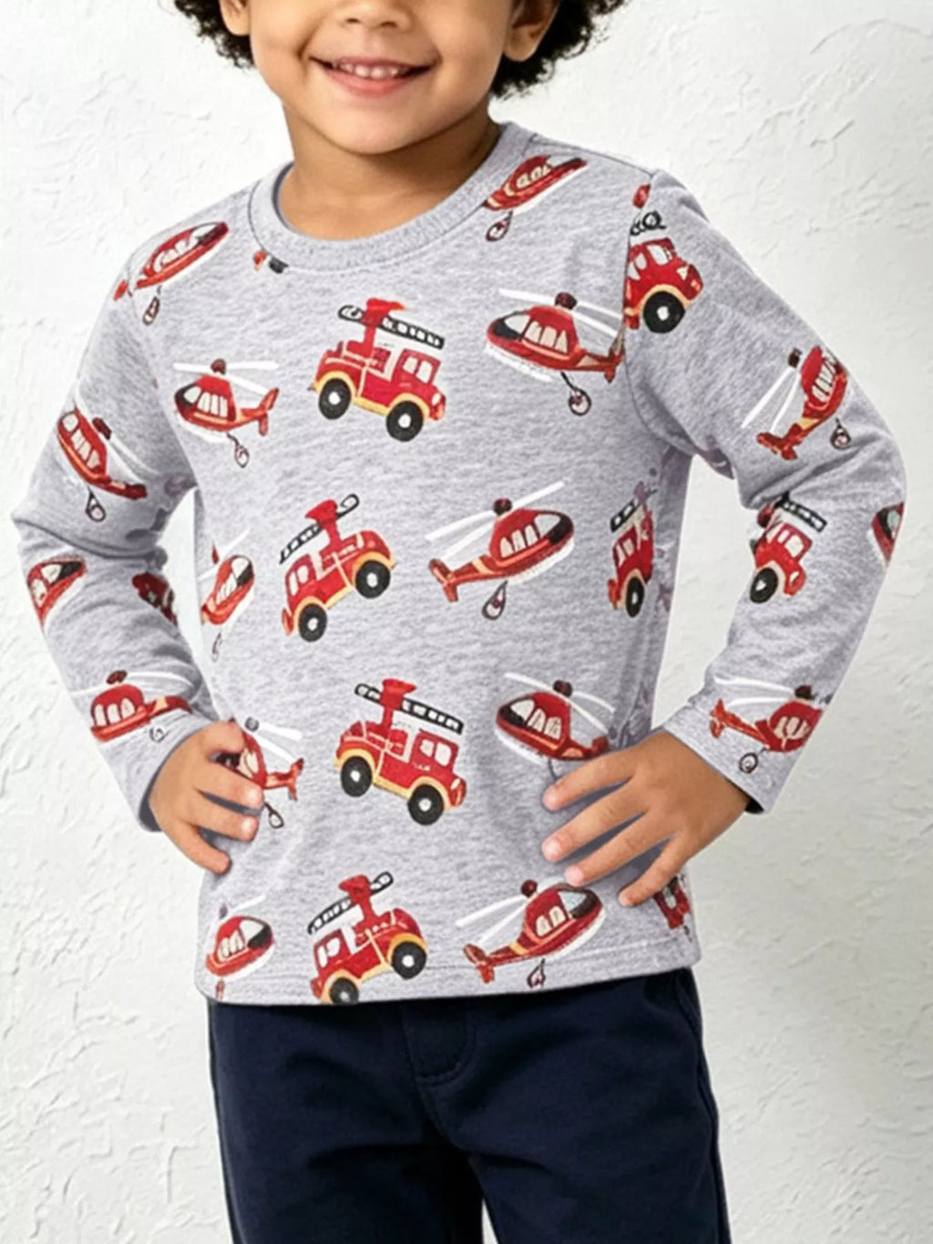 Child wearing a gray long-sleeve top printed with red fire trucks and helicopters.