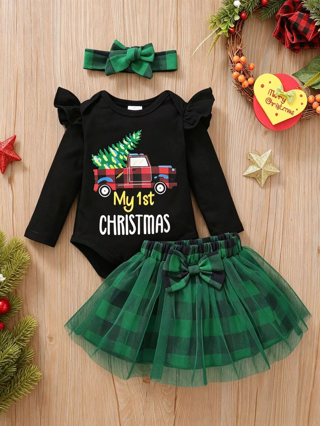 Flat lay of a black My 1st Christmas ruffle-shoulder bodysuit, green plaid tulle skirt and matching bow headband.
