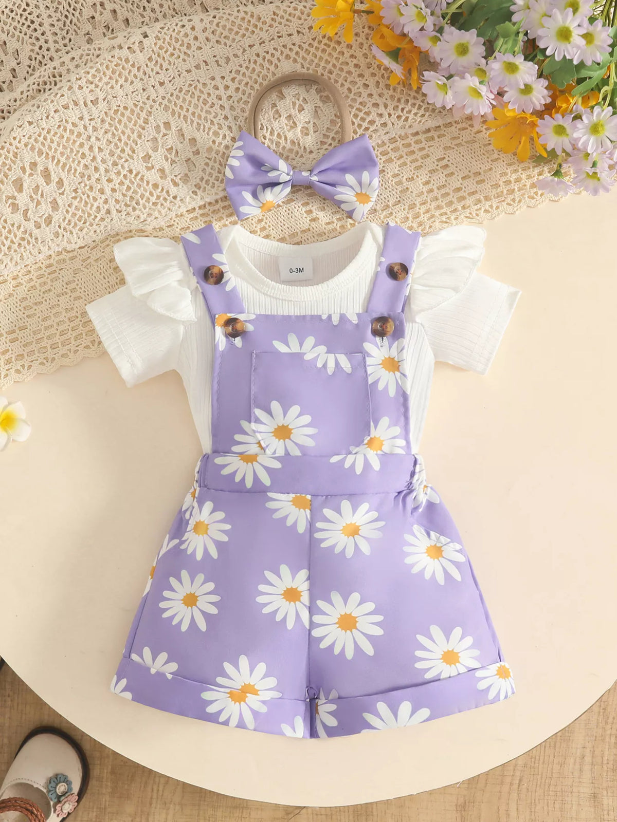 White baby bodysuit with lavender daisy-print short overalls and matching bow headband.