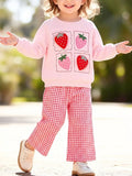Child wearing a pink strawberry graphic sweatshirt and pink gingham flared pants outdoors.