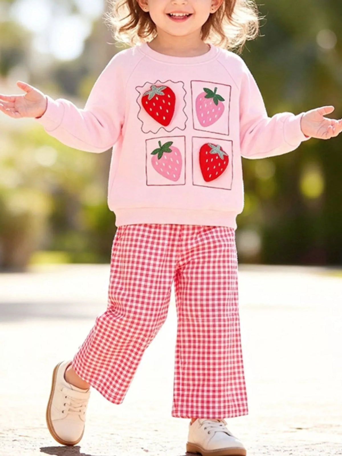 Child wearing a pink strawberry graphic sweatshirt and pink gingham flared pants outdoors.