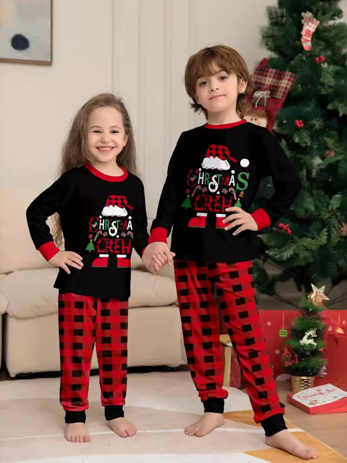 Two children wearing matching black Christmas graphic tops and red plaid pants.