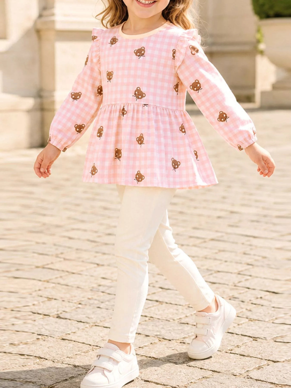 Child wearing a pink gingham bear-print ruffle top with cream leggings.