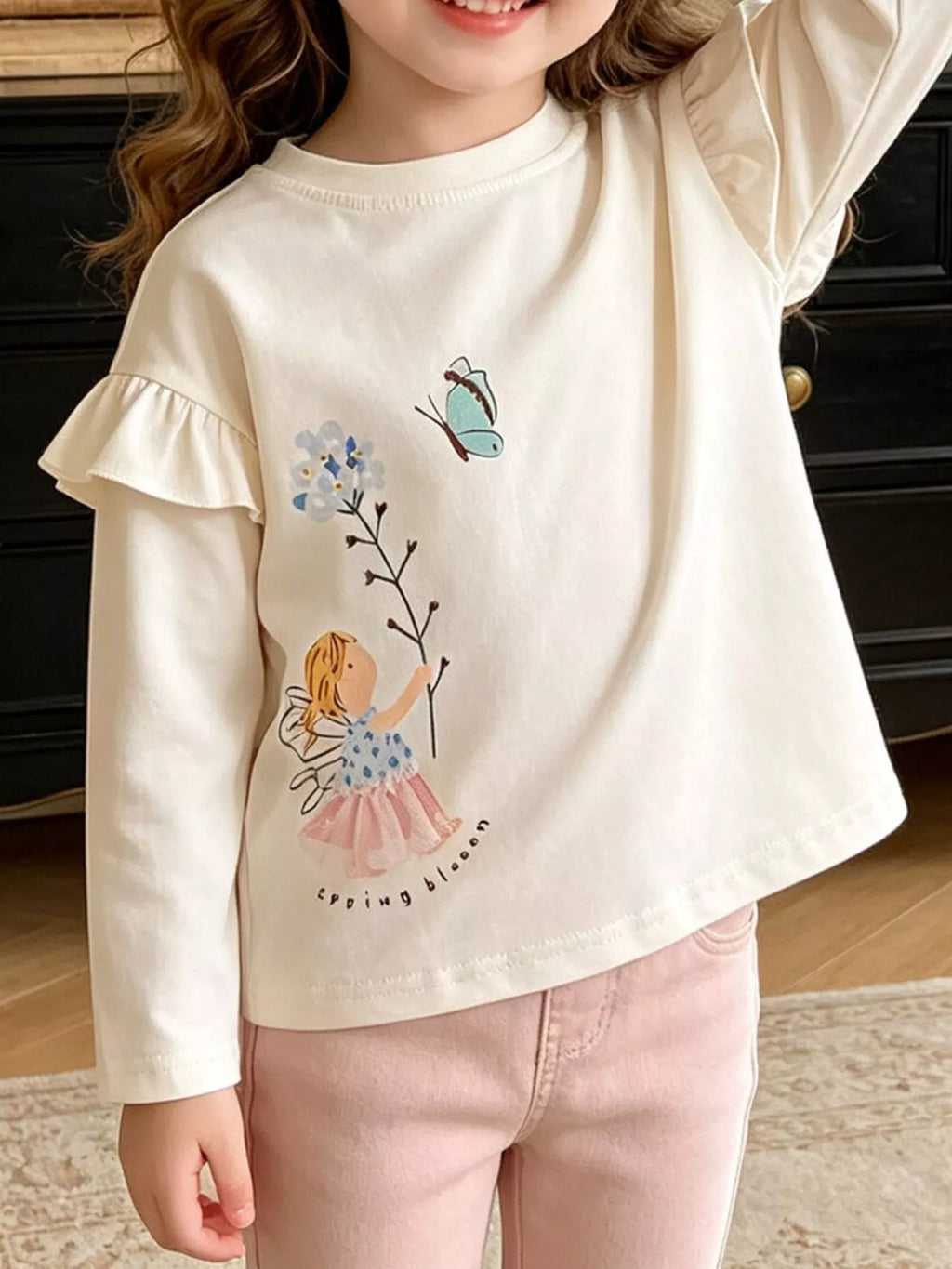 Child wearing a cream ruffle-shoulder top with fairy and butterfly artwork.