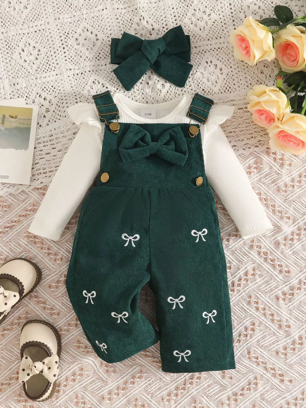 White ribbed baby bodysuit with dark green bow-embroidered overalls and matching headband displayed flat