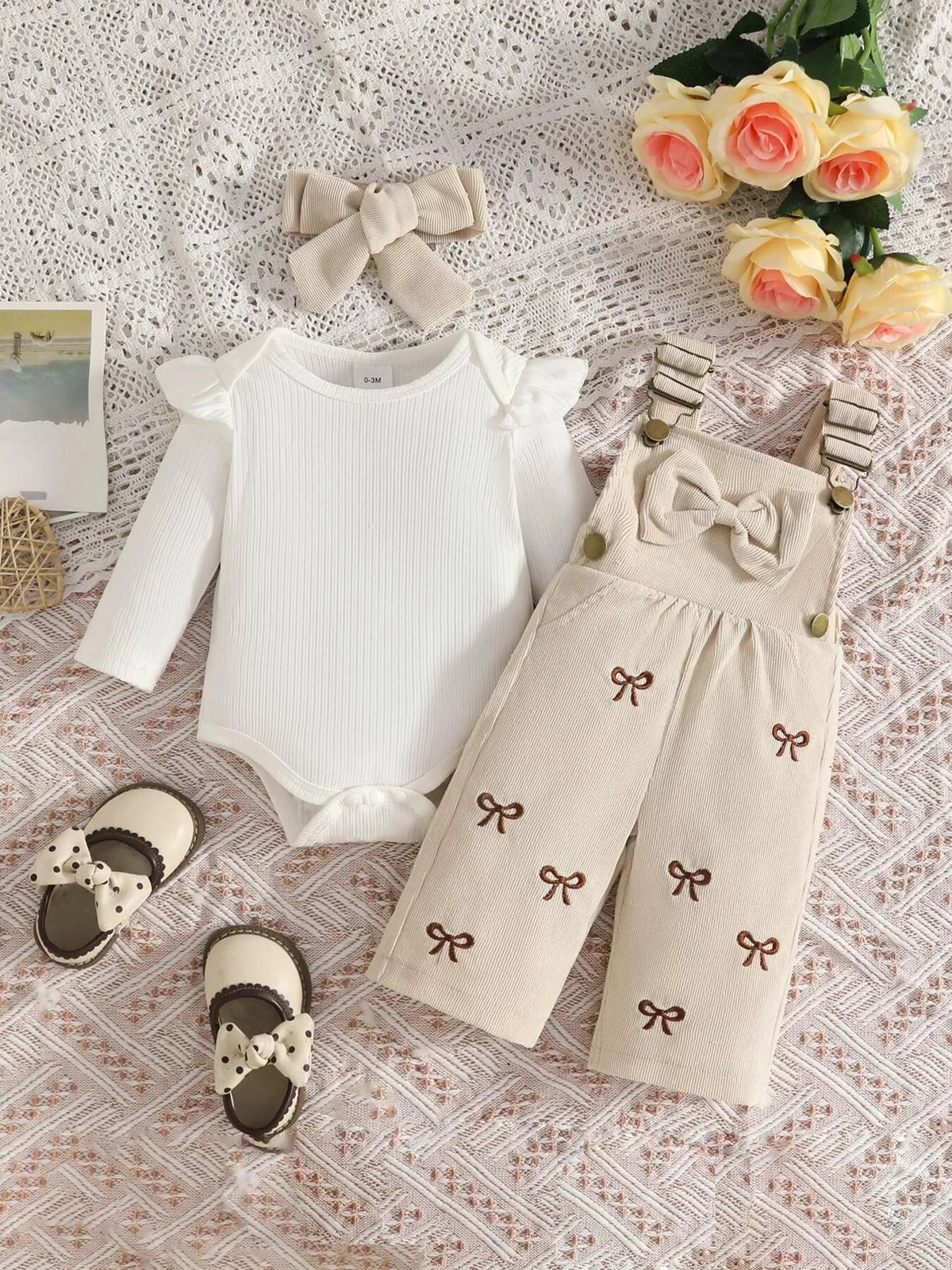 White ribbed baby bodysuit with beige bow-embroidered overalls and matching headband displayed flat