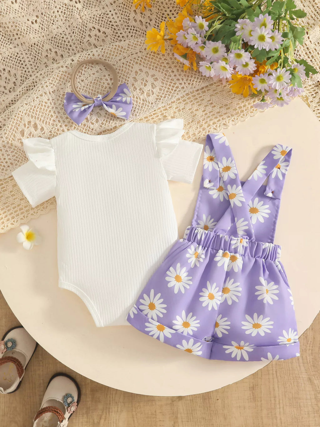 Back flat lay of the white bodysuit, lavender daisy-print short overalls and matching bow.