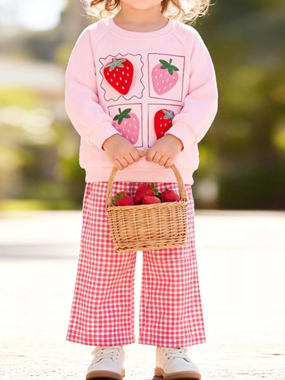 Child modeling the pink strawberry sweatshirt and gingham pants while holding a basket.